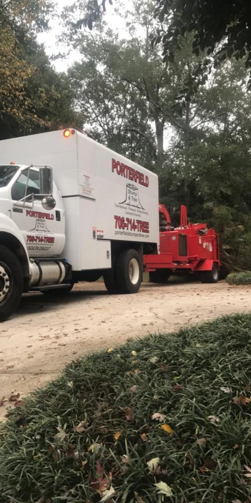 A white Porterfield Stump & Tree service truck parked on a paved driveway, towing a large red commercial wood chipper. The truck displays the company logo and a 706 area code phone number against a backdrop of lush green trees.