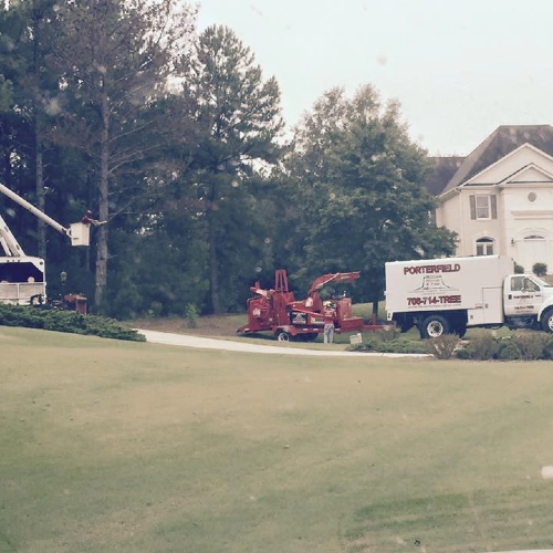 A Porterfield Stump & Tree service truck and wood chipper parked on a green lawn in front of a large house, with a bucket lift extended for tree work.
